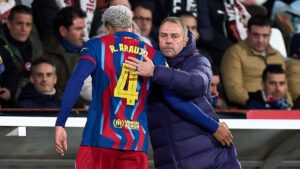 Hansi Flick steht nachdenklich an der Seitenlinie des FC Barcelona, im Hintergrund trainiert Ronald Araujo.