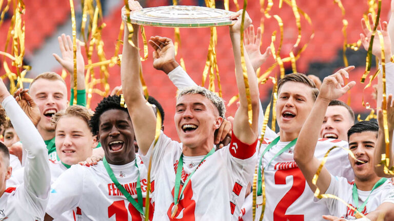 Symbolbild: Junge Fußballspieler im Zweikampf auf dem Rasen, im Hintergrund ein Stadion
