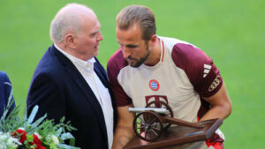 Harry Kane im Trikot des FC Bayern München jubelt über ein Tor, während Uli Hoeneß auf der Tribüne applaudiert