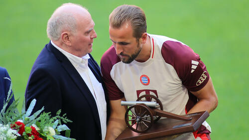 Harry Kane im Trikot des FC Bayern München jubelt über ein Tor, während Uli Hoeneß auf der Tribüne applaudiert