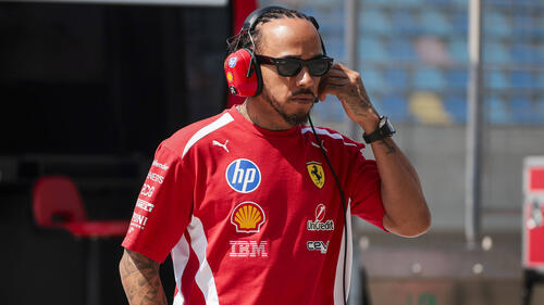 Lewis Hamilton im roten Ferrari-Rennanzug blickt entschlossen in die Ferne, im Hintergrund unscharf die Boxengasse.