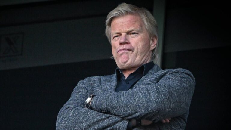 Bayern-Präsident Herbert Hainer blickt ernst, während im Hintergrund Jamal Musiala im Trikot des FC Bayern München spielt.