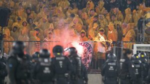 Polizei sichert das Spielfeld im Stadion ab, während im Hintergrund Pyrotechnik und Bengalos im Fanblock brennen.