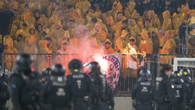 Polizei sichert das Spielfeld im Stadion ab, während im Hintergrund Pyrotechnik und Bengalos im Fanblock brennen.
