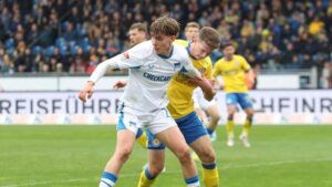 Kennet Eichhorn im Trikot von Hertha BSC auf dem Spielfeld, im Hintergrund die Vereinswappen von Liverpool, Bayern und BVB.